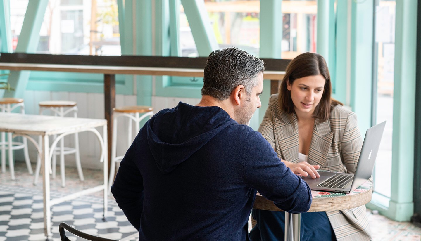 two people sitting in a cafe looking at a laptop on a table